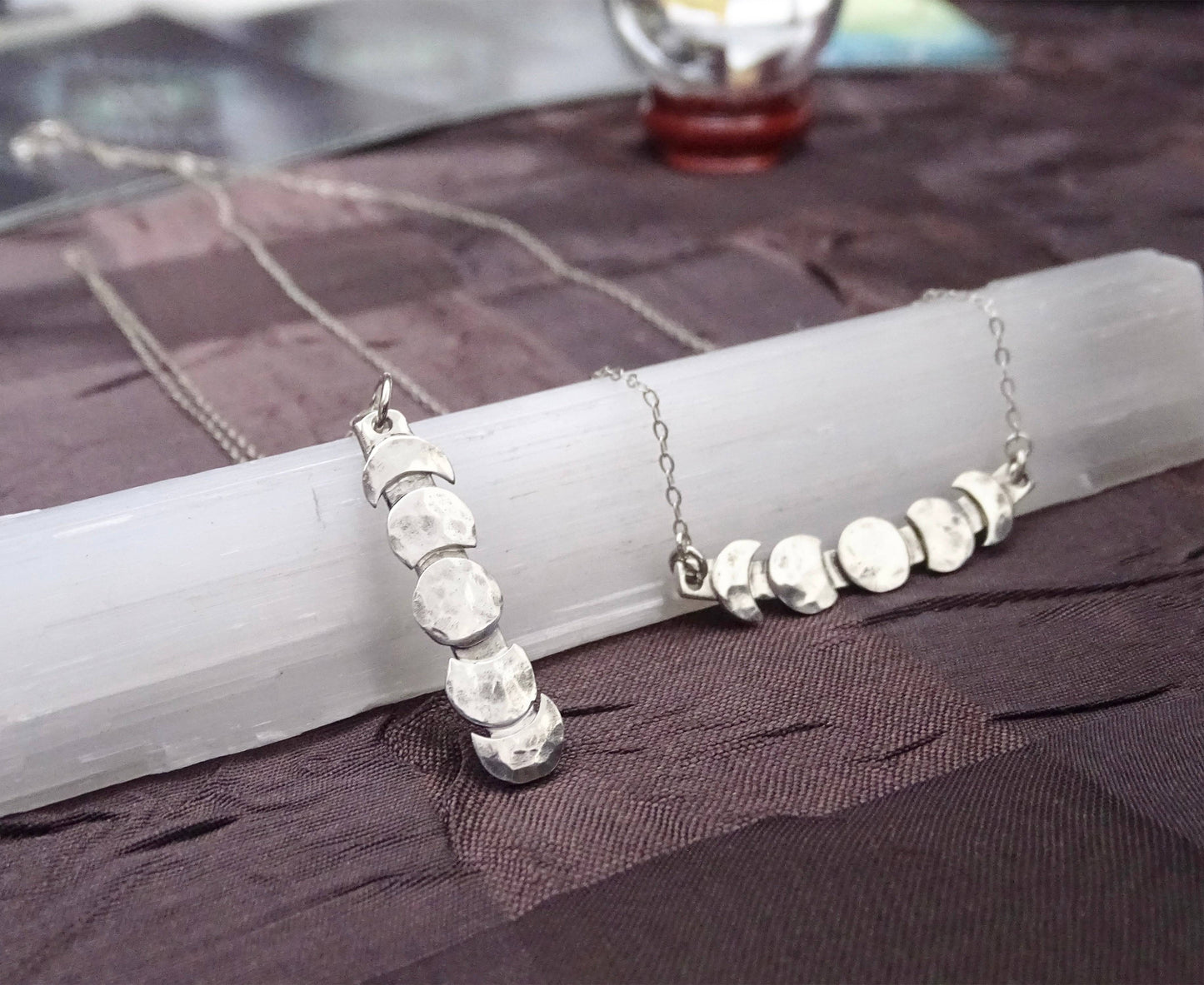 Vertical and horizontal moon phase necklaces displayed on selenite wand, featuring hammered sterling silver lunar designs.