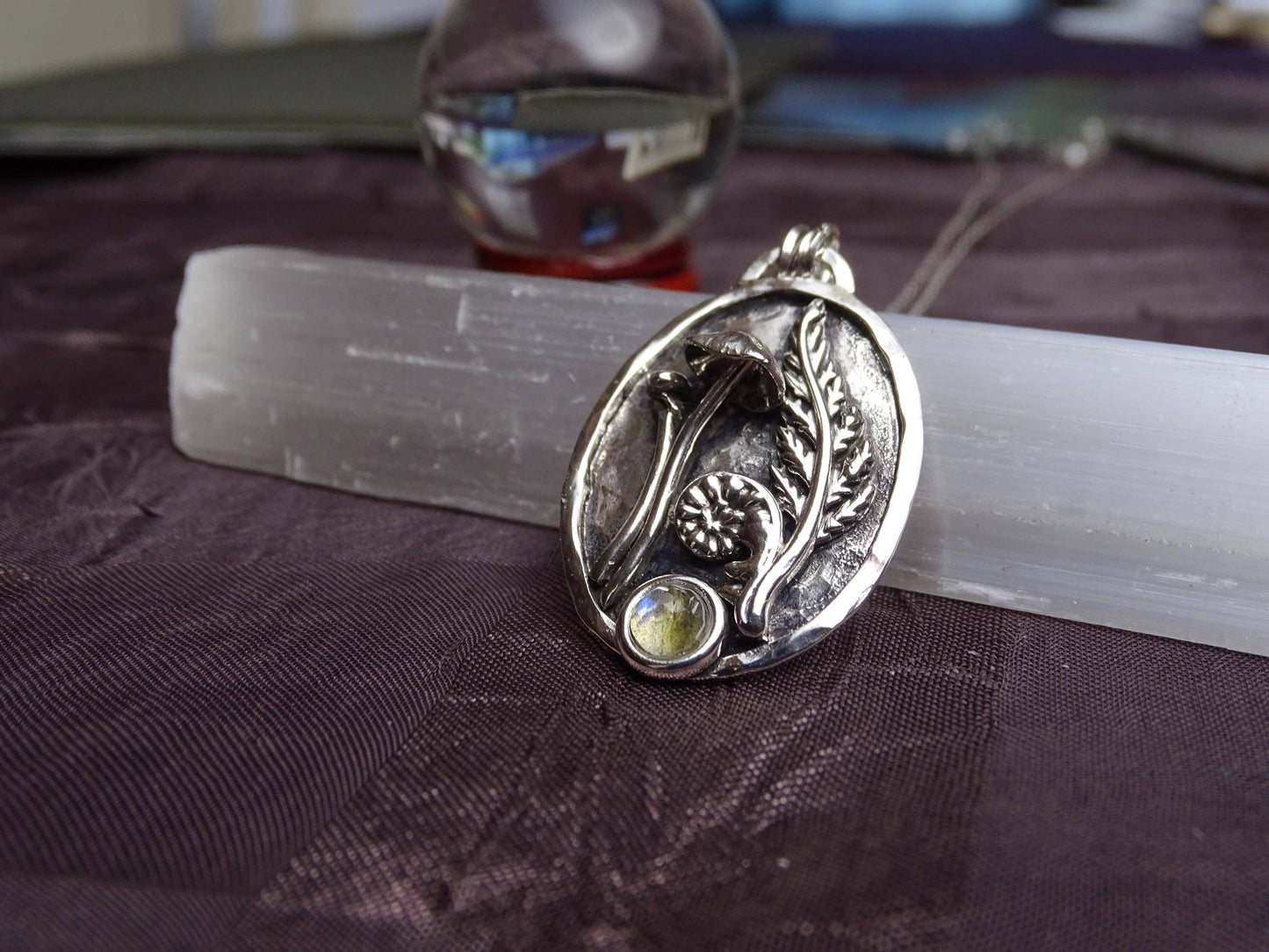 Artisan sterling silver pendant with mushroom, fern, and labradorite gemstone, displayed on a white selenite wand over a dark fabric background.