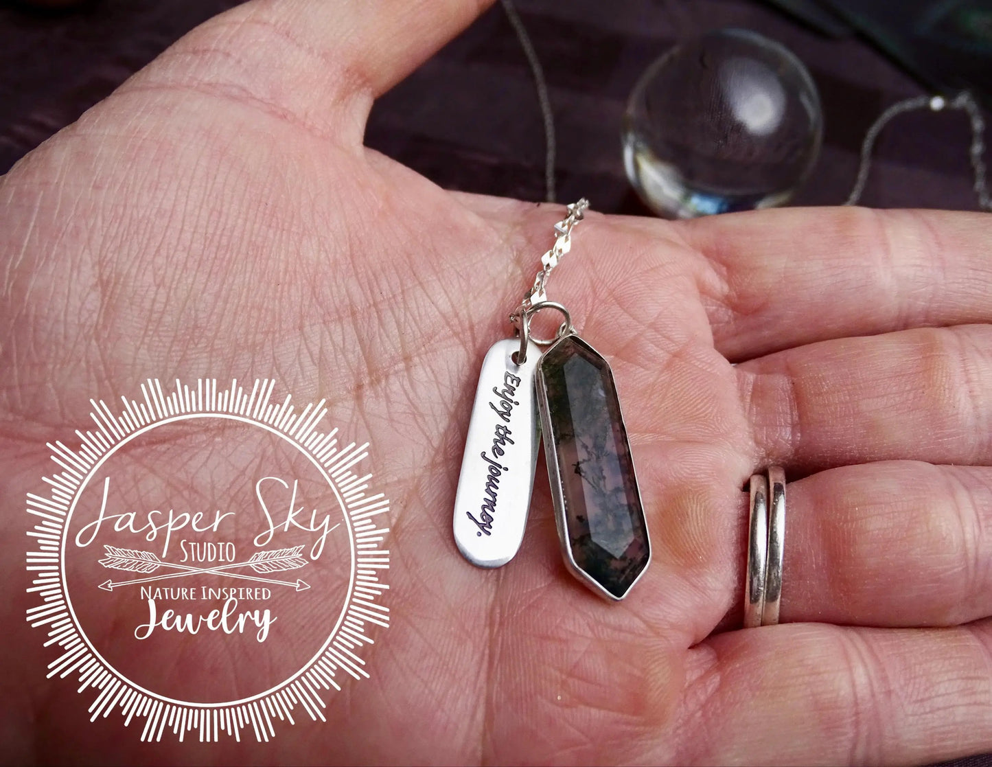 Hand holding sterling silver moss agate hexagon pendant with 'Enjoy the Journey' charm, highlighting its size and unique design.